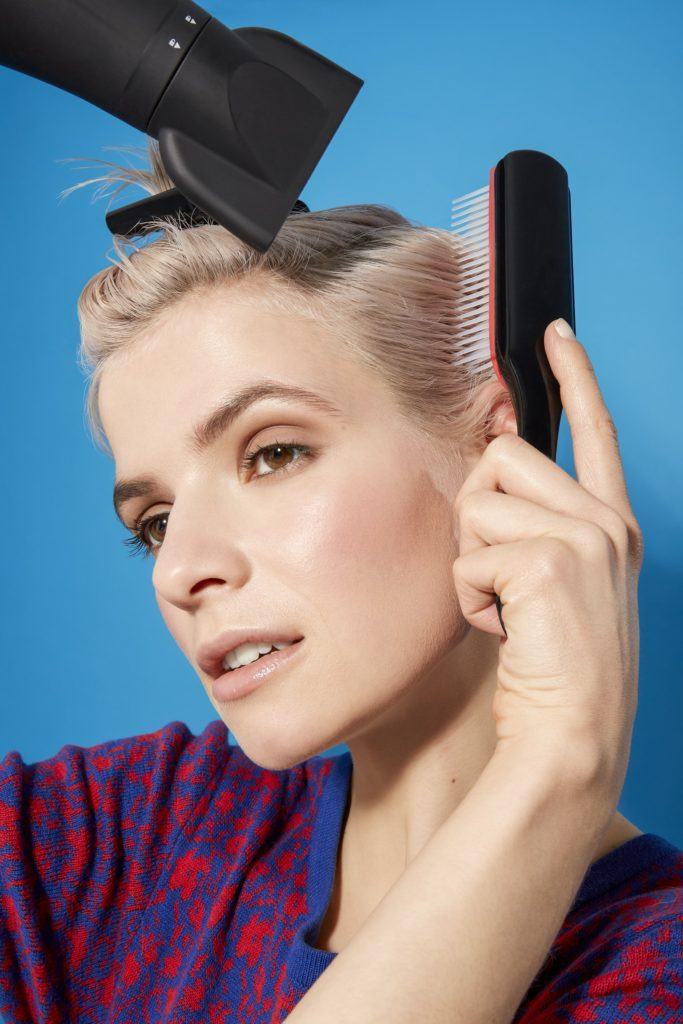 woman with platinum blonde hair in a pixie cut style using a brush and hairdryer to style her hair