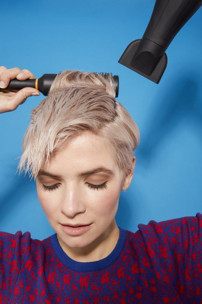 woman with platinum blonde hair in a pixie cut style, looking down using a brush and hairdryer to style her hair