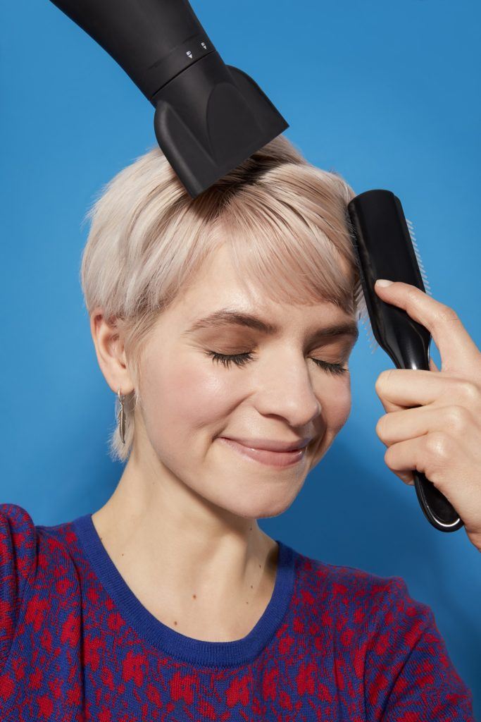 woman with platinum blonde hair in a pixie cut style using a brush and hairdryer to style her hair