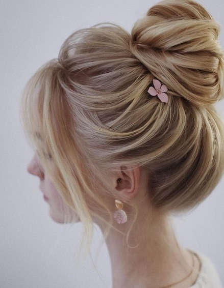 Wedding updos: Side view of a blonde woman with her hair in a high twisted bun with a long fringe and a pink flower hair slide