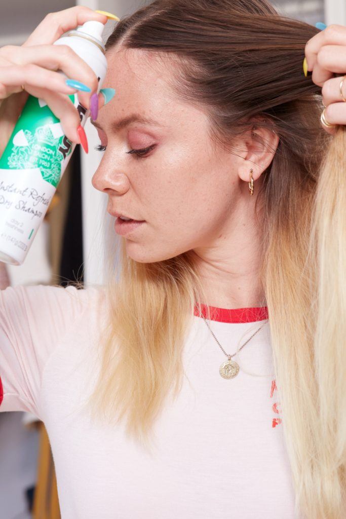 heart braid tutorial: close up shot of woman spraying dry shampoo into her roots, wearing white top and necklace, posing in a bedroom