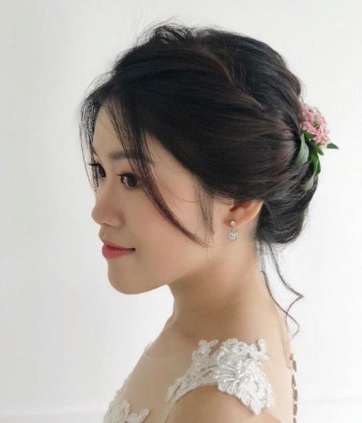 Wedding updos: Side profile of an Asian woman with brunette hair styled in a side twisted updo with loose strands, with a flower hair accesory