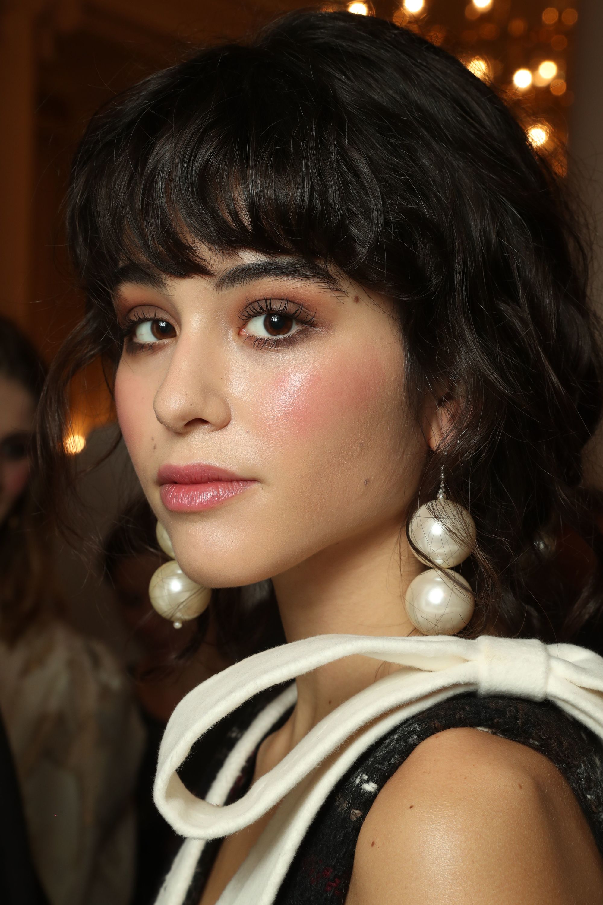 Haircuts for thick hair: Close-up backstage image of a model with dark brunette bowl cut hair with oversized pearl earrings.