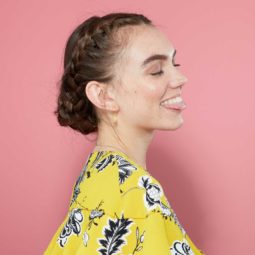 side view of a brunette model sticking her tongue out with her hair in a french braided bun updo wearing a yellow floral dress against a pink background