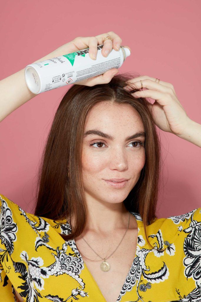 brunette model spraying toni & guy dry shampoo into her roots wearing a yellow floral dress