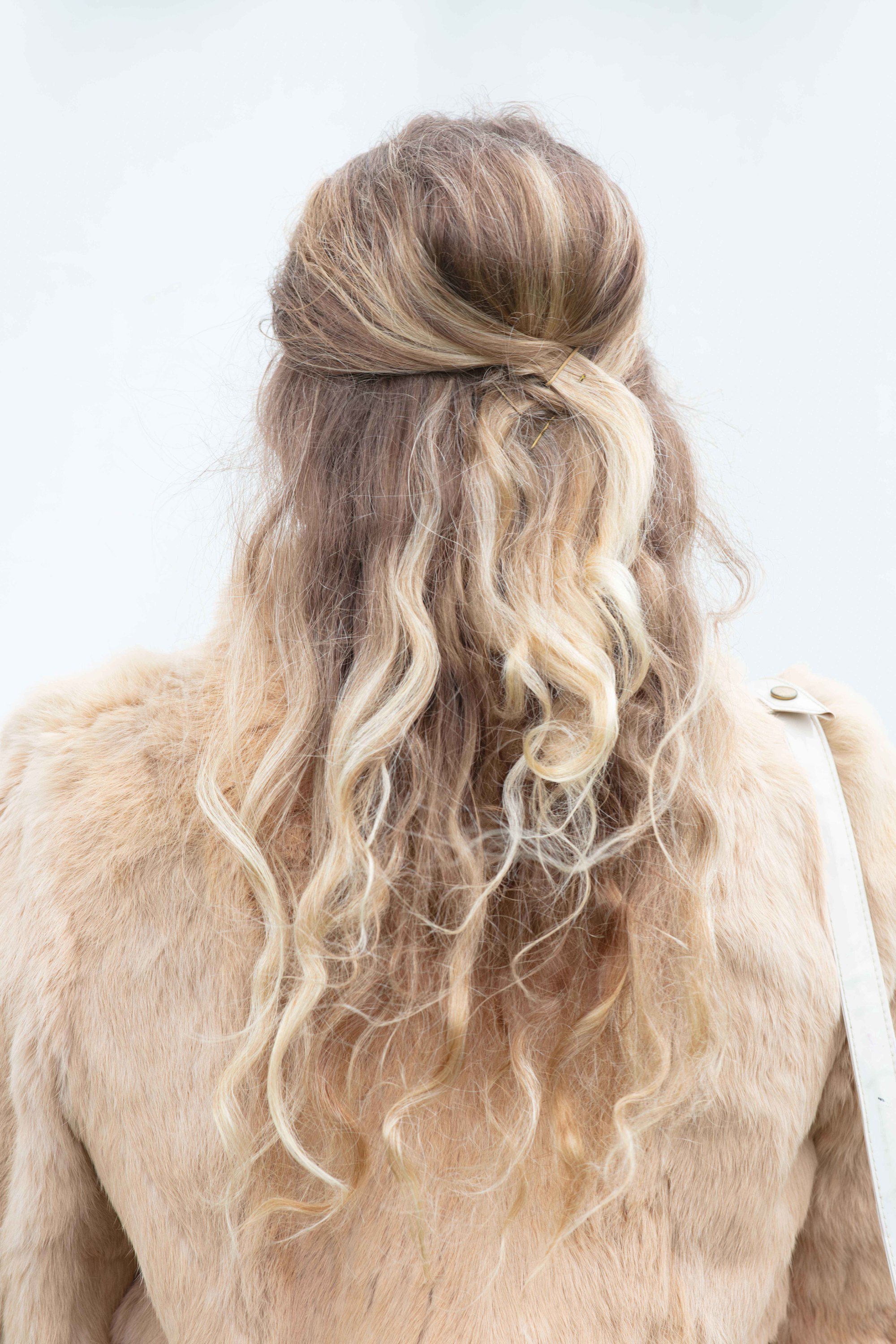 Retro hairstyles: Close up back shot of a woman with long bronde hair styled into a half-up, half-down beehive, wearing a furry jacket and posing at the Goodwood festival