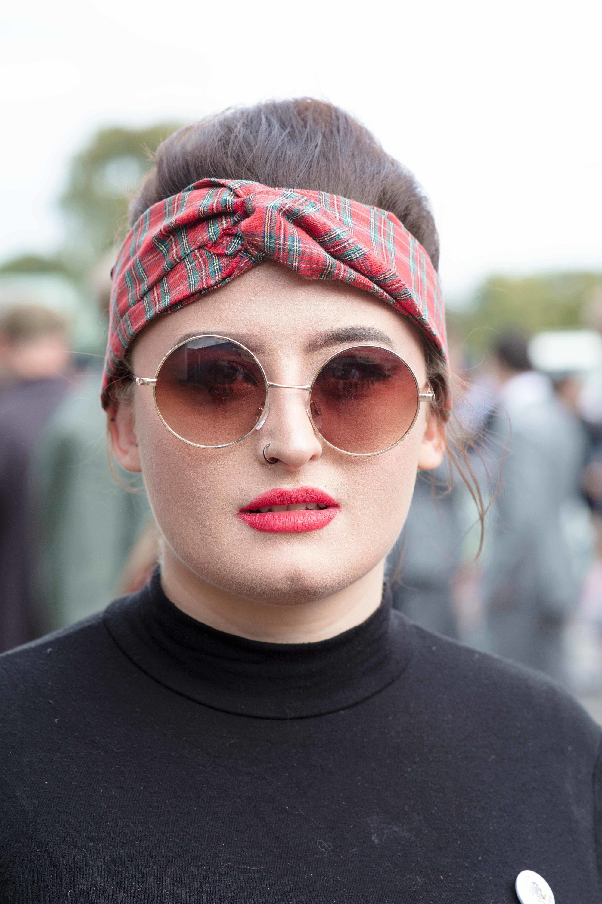 Retro hairstyles: Close up shot of a woman with dark brown hair styled into a beehive updo with a headwrap, wearing black and John Lennon style glasses at Goodwood festival