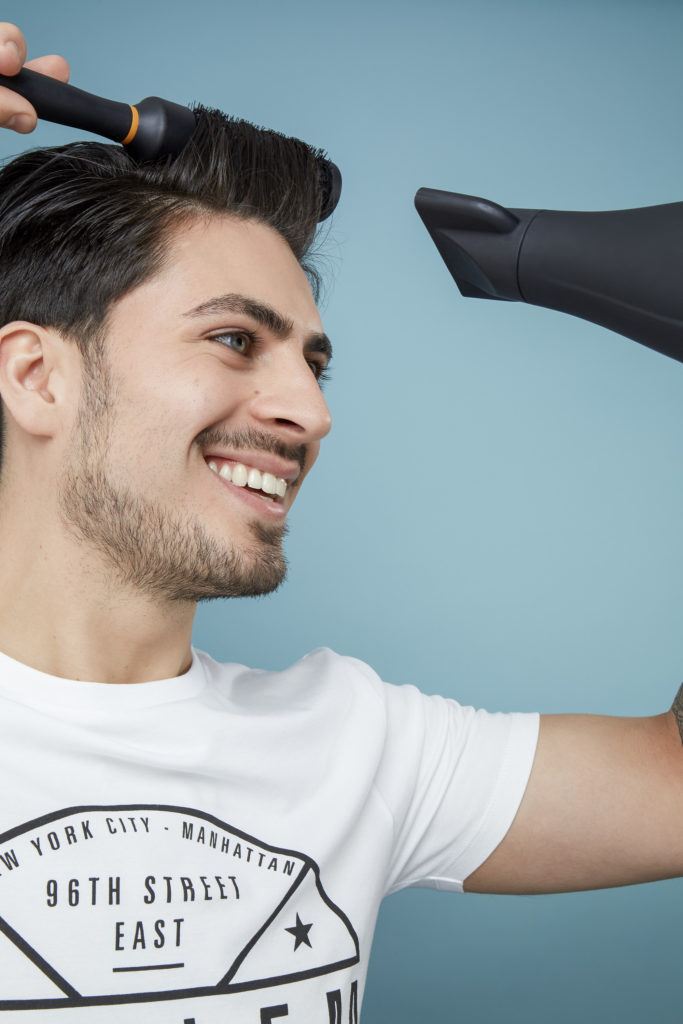 How to blow dry men's hair: Brown haired man using a round brush to blow dry his hair, wearing a white t-shirt