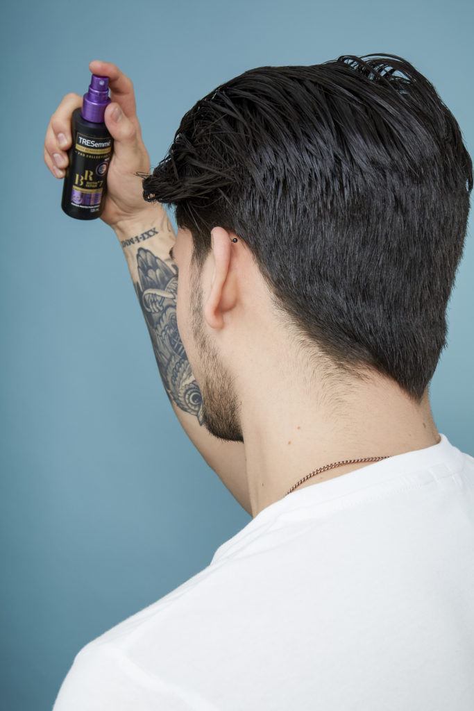 How to blow dry men's hair: Back view of a brown haired man with wet hair spraying Tresemme primer on his hair, wearing a white t-shirt
