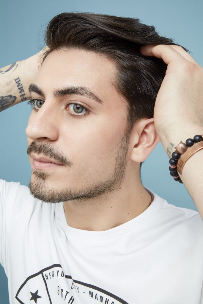 How to blow dry men's hair: Man with brown swept back hair and facial hair styling his hair with his fingers wearing a white tshirt