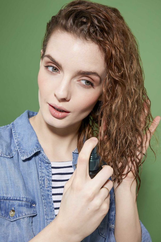 How to scrunch hair tutorial: Woman with brown curly hair spraying leave in conditioner into her hair, which is swept over one shoulder