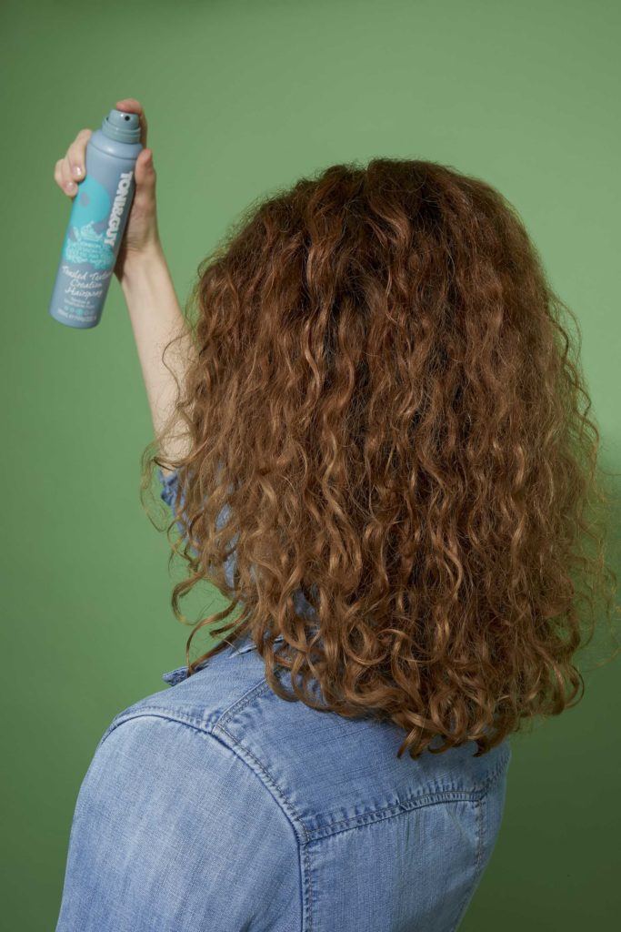 How to scrunch hair tutorial: Back view of a woman with shoulder length brown curls spraying her hair with TONI&GUY hairspray