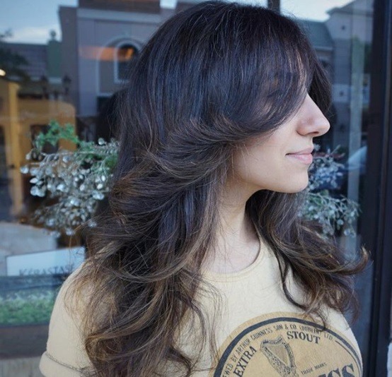 Long shag haircut: Close up shot of a woman with dark brown long shaggy hair with feathered layers, wearing slogan top and posing outside a salon