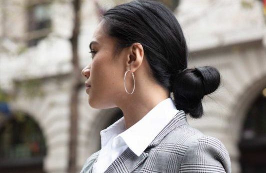 Knot bun hairstyle: Close up shot of a woman with a dark low knot bun wearing checked jacket with white top, wearing belt bag and posing on the street
