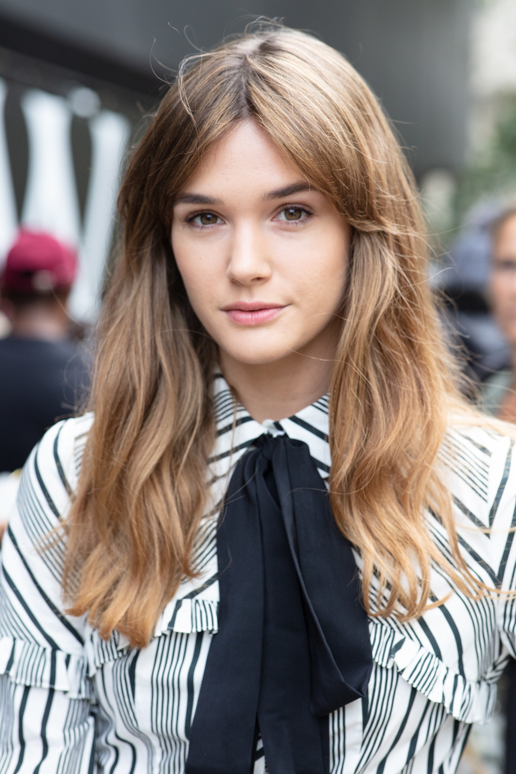 Long shag haircut: Close up shot of actress Sai Bennett with a chestnut brown hairstyle with bardot bangs and loose shaggy waves, wearing a pussy bow shirt and posing on the street