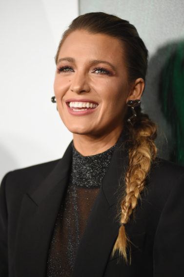 Winter hairstyles 2018: Blake Lively with her dark blonde hair in a textured side braid, wearing a black blazer and sheer black high neck top