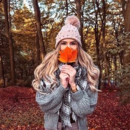 Bonfire hairstyles: Young woman in the woods with long wavy blonde hair, wearing grey jacket with scarf and pink bobble hat, covering her face with a fallen leaf