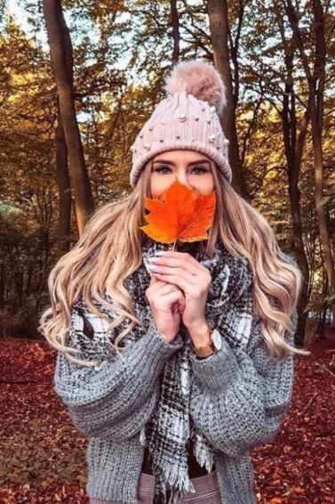 Bonfire hairstyles: Young woman in the woods with long wavy blonde hair, wearing grey jacket with scarf and pink bobble hat, covering her face with a fallen leaf