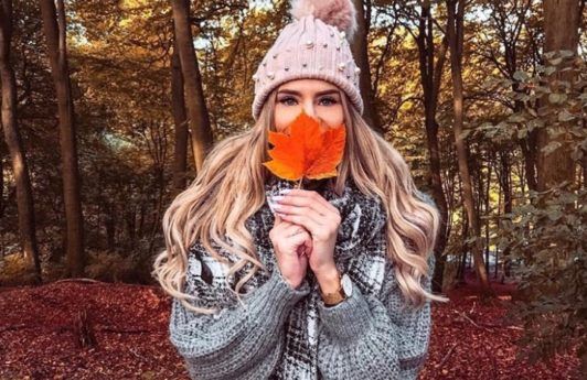Bonfire hairstyles: Young woman in the woods with long wavy blonde hair, wearing grey jacket with scarf and pink bobble hat, covering her face with a fallen leaf