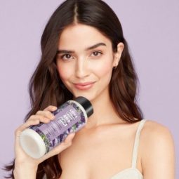 Photo of a brunette model with long waves, holding a bottle of Love Beauty And Planet against a lilac purple background