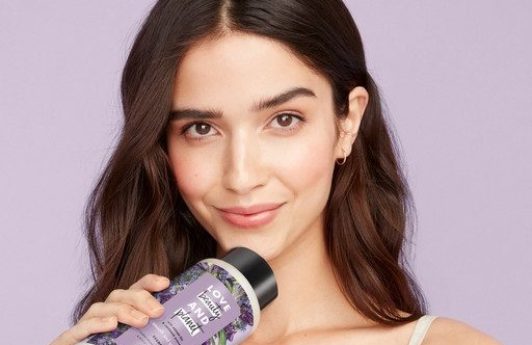 Photo of a brunette model with long waves, holding a bottle of Love Beauty And Planet against a lilac purple background