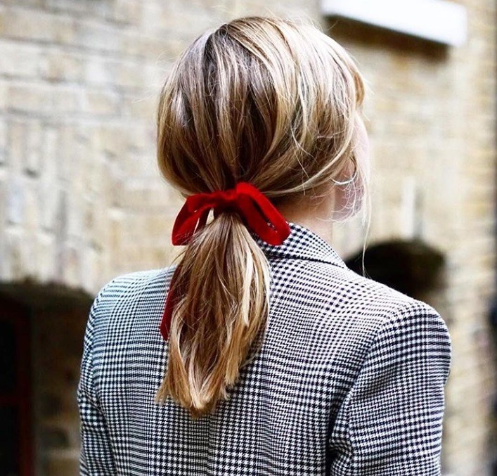 Bonfire hairstyles: Back shot of woman with bronde medium length hair styled into a low ponytail and tied up with a red bow, wearing a checked blazer and posing outside