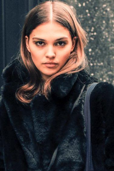 Winter hair mask guide: Street shot of young woman with chestnut brown hair tucked into a big, black fake fur jacket, wearing a tote black and a cross body handbag while posing