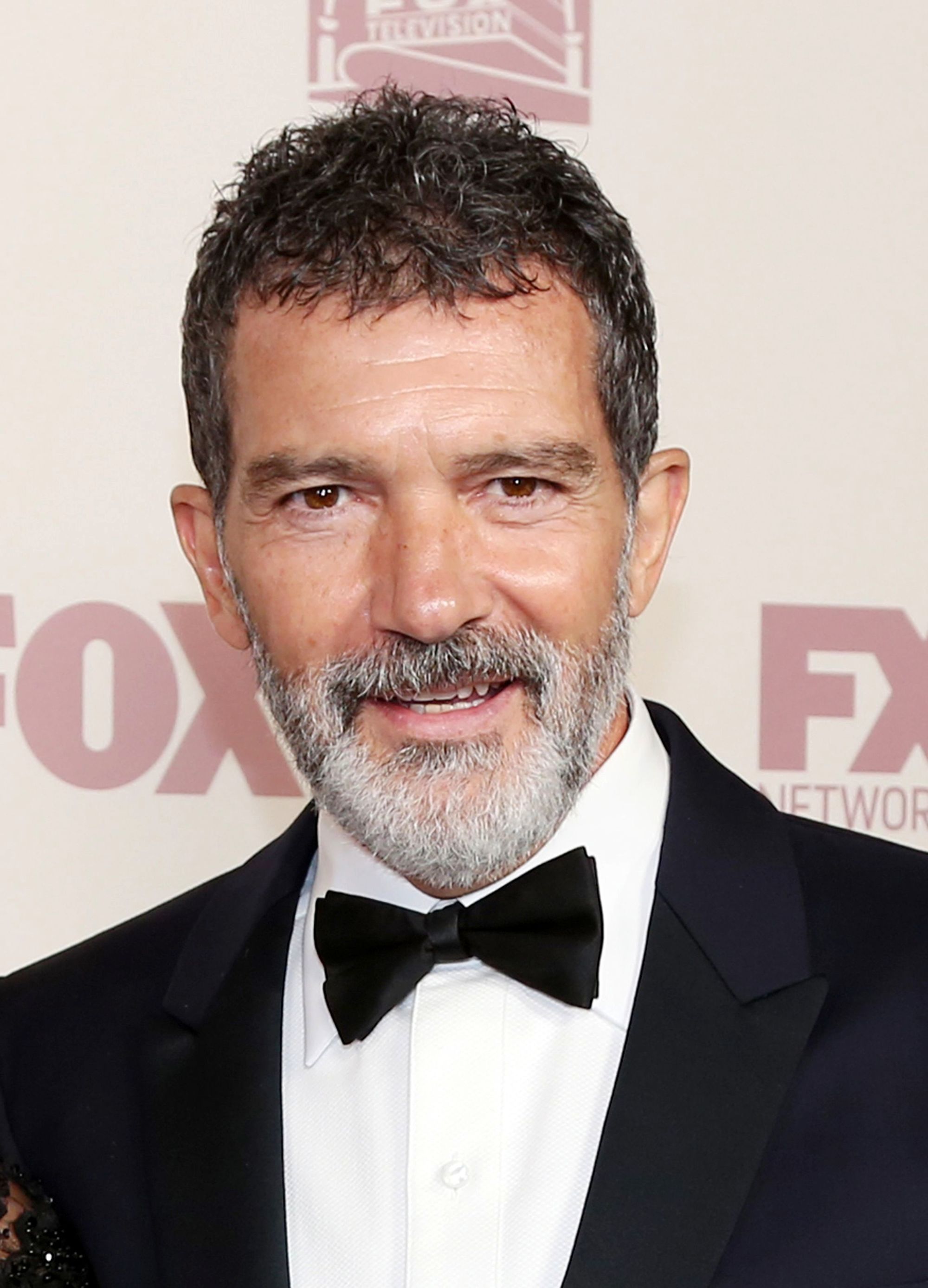 Hairstyles for men over 50: Antonio Banderas with brown French crop hairstyle with salt and pepper grey beard wearing a smart suit.