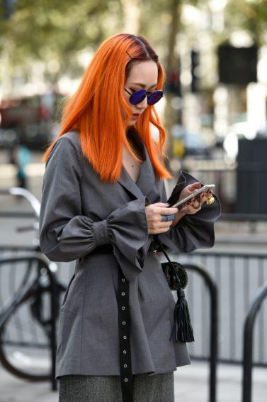 Woman wearing grey jacket with long orange hair, holding phone and posing on the street