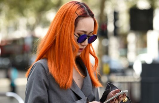 Woman wearing grey jacket with long orange hair, holding phone and posing on the street