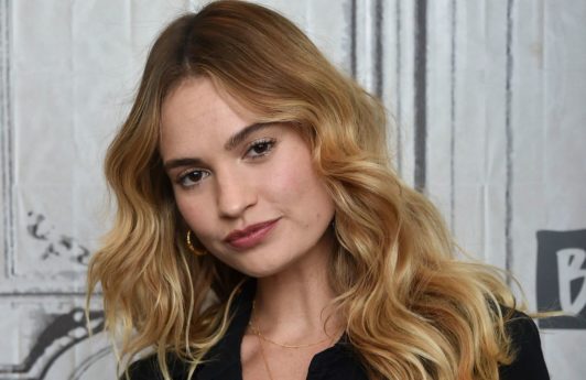 How to use volume spray guide: Lily James with bronde long wavy, voluminous hair, wearing black boiler suit and posing on the red carpet
