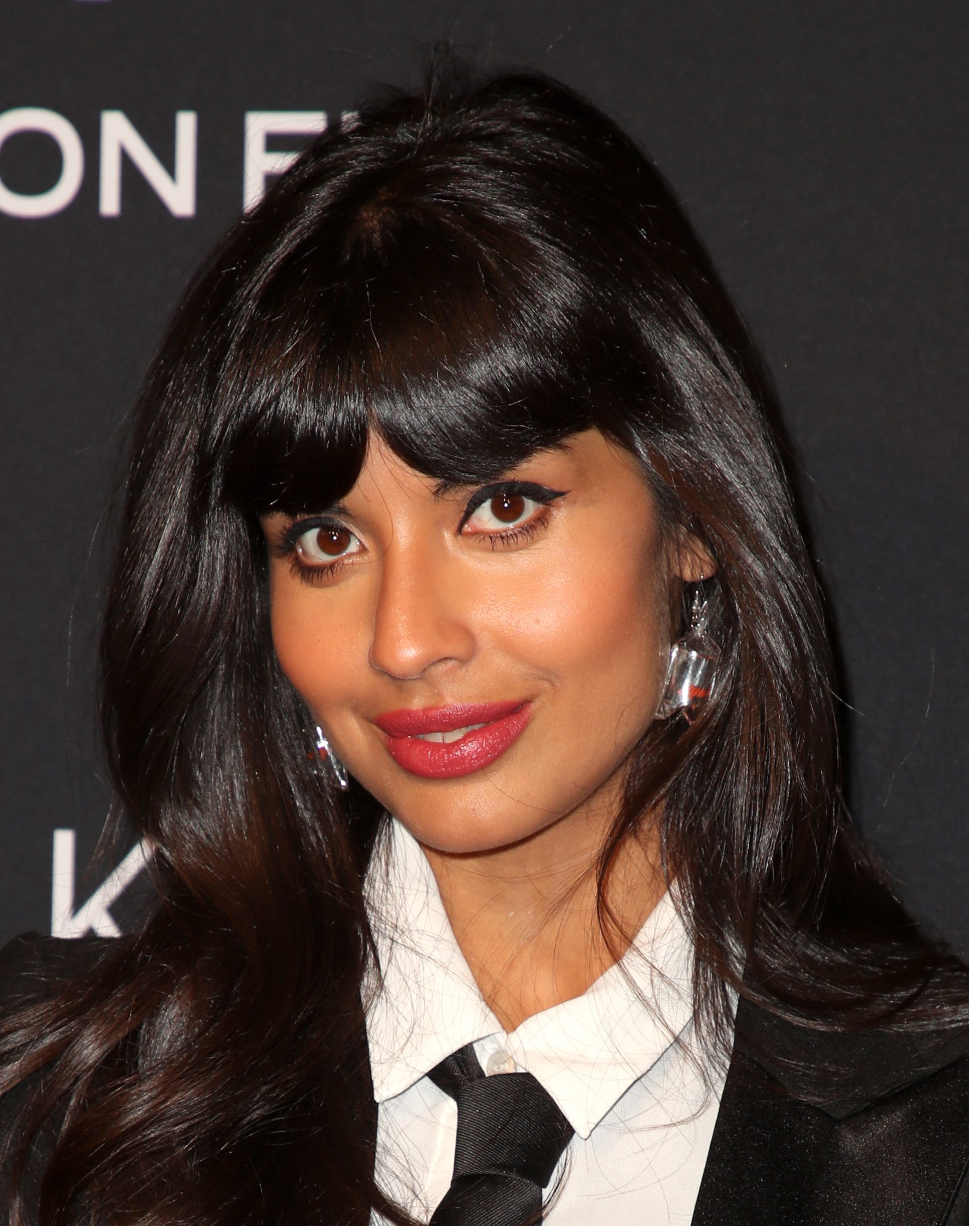Bollywood hair: Jameela Jamil with dark chocolate brown hair and thick, heavy bangs, wearing white top and jacket on the red carpet