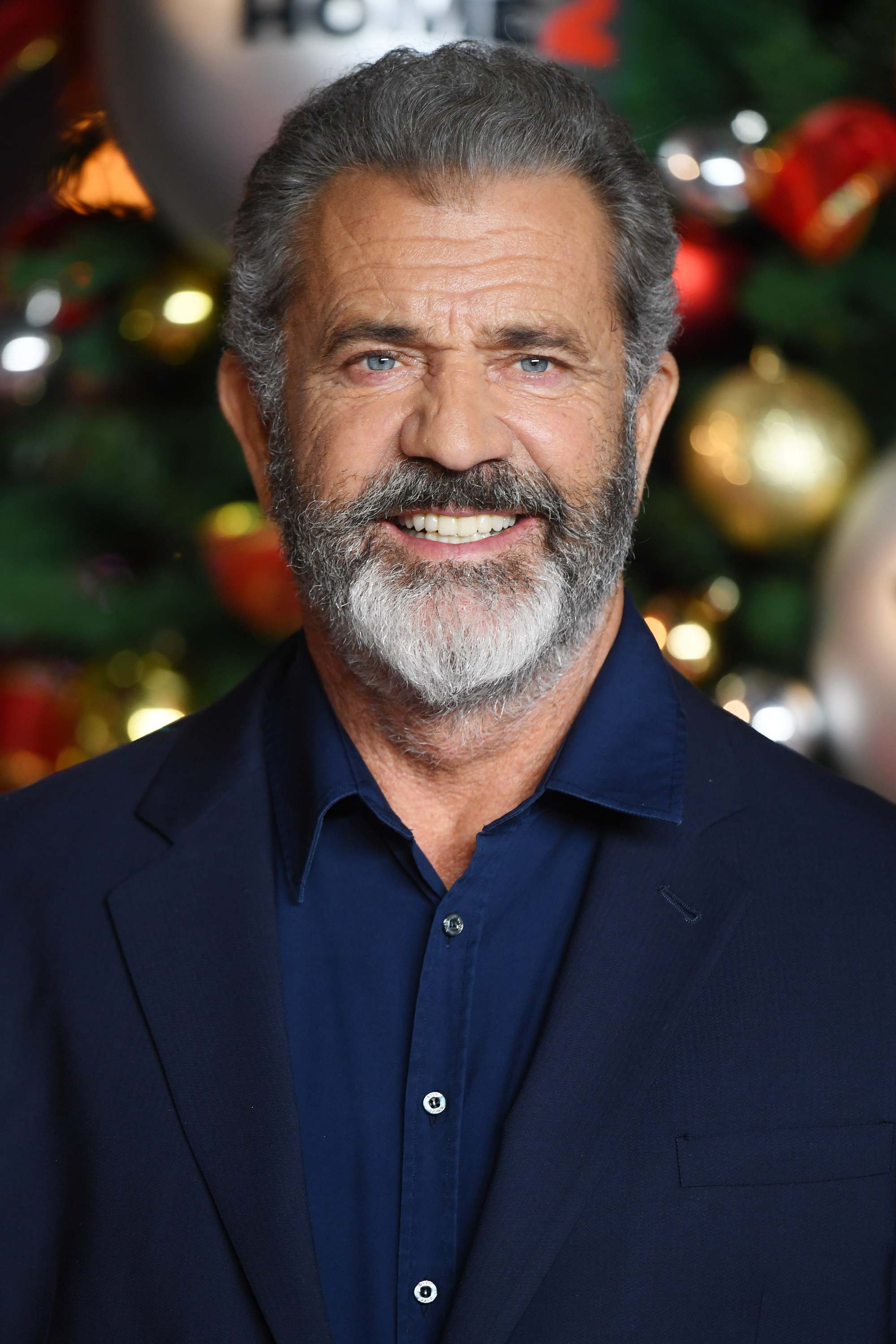 Hairstyles for men over 50: Mel Gibson with salt and pepper hair and full beard wearing a relaxed suit in front of a christmas tree.