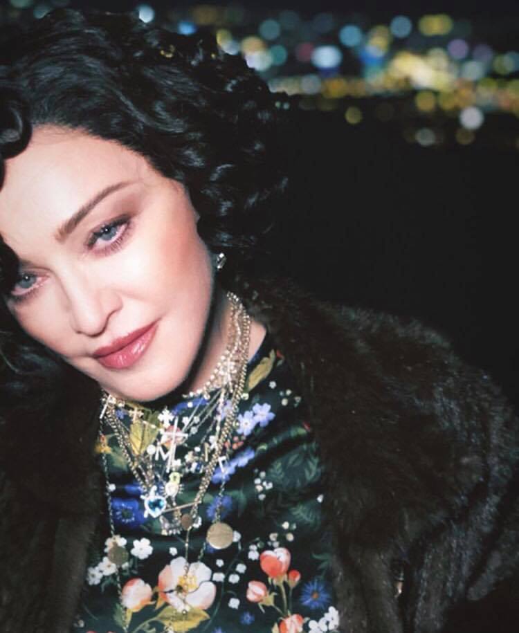 Close up shot of Madonna with dark hair, with retro curls, wearing a floral top and fur coat with necklaces posing for photo
