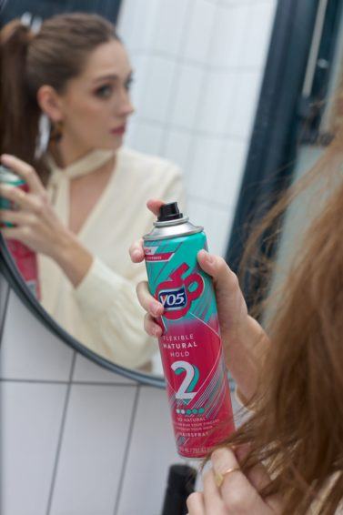 Woman spraying her brown hair with VO5 Flexible Hold Hairspray in the mirror.
