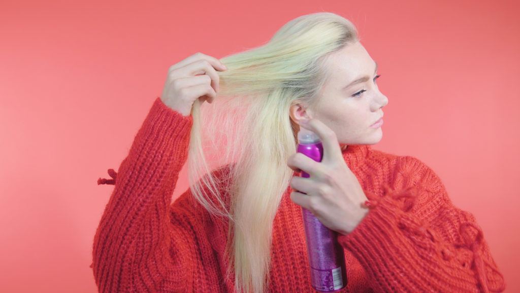Side Dutch braid tutorial: Blonde girl with long straight hair spraying volumising spray into her roots, wearing a red knit jumper