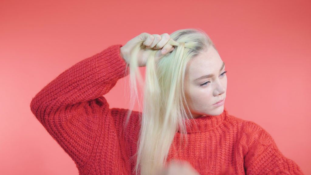 Side Dutch braid tutorial: Blonde girl with long straight hair dividing a section of hair into 3 to braid, wearing a red knit jumper
