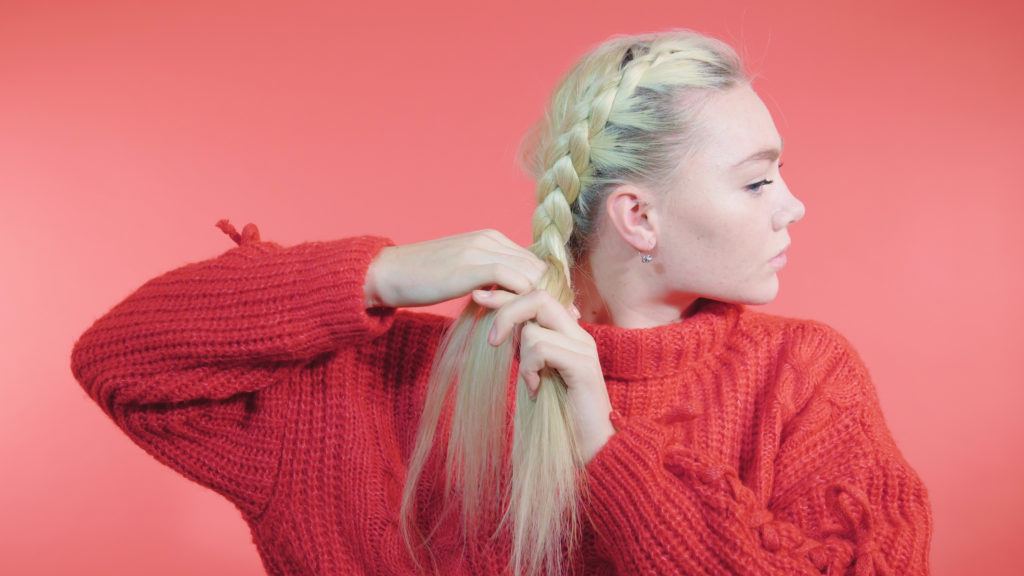 Side Dutch braid tutorial: Blonde girl braiding her hair into a side Dutch braid, wearing a red knit jumper