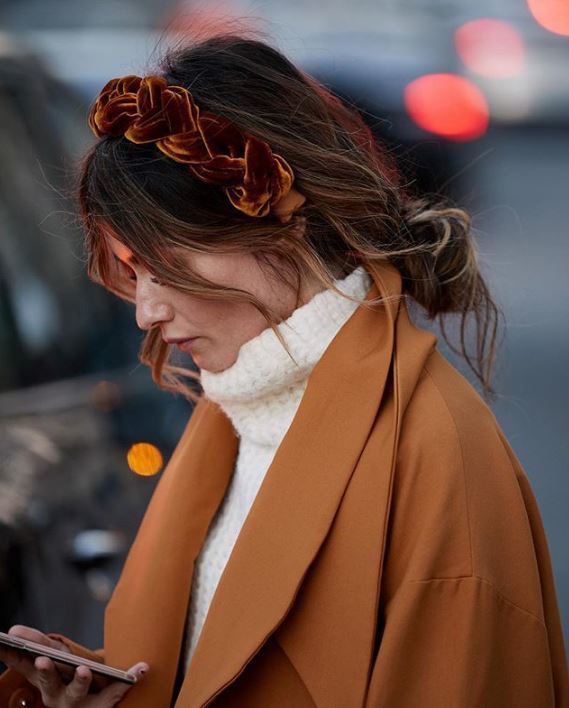 Bank holiday hairstyles: Woman with brown balayage wavy hair in messy low bun wearing a plaited headband and winter outfit.