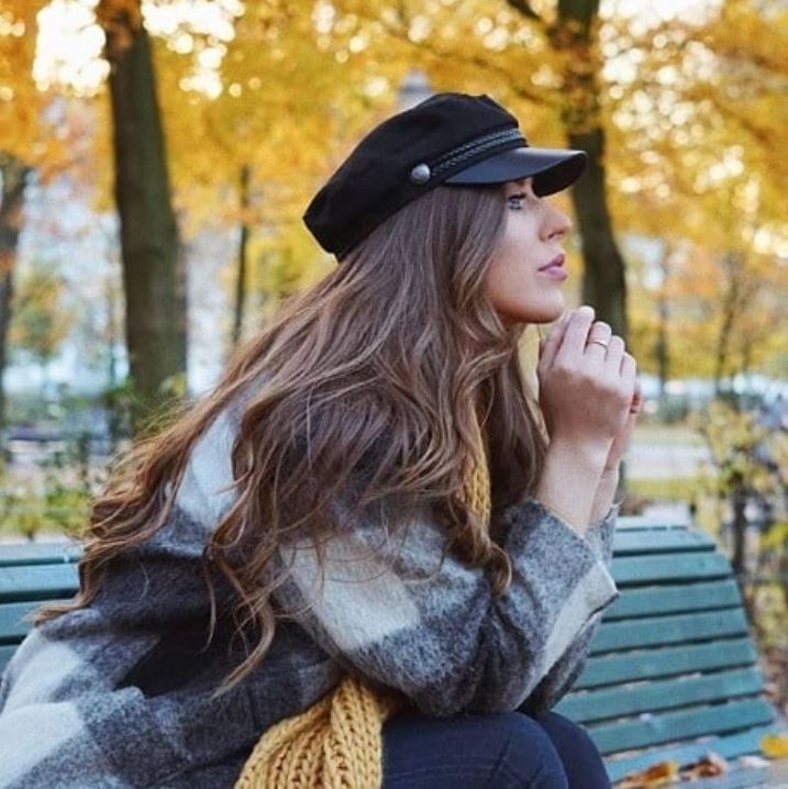 Bank holiday hairstyles: Woman with long wavy brown hair wearing a baker boy hat sitting on a bench.