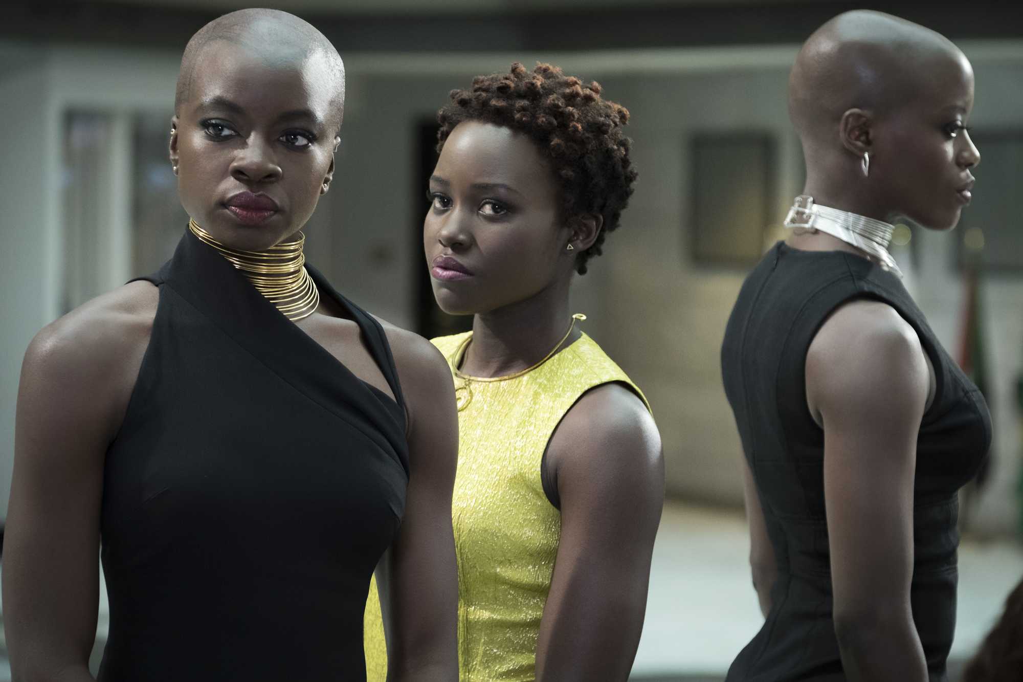 Marvel hairstyles: Danai Gurira in Black Panther with shaved head wearing a black dress standing with Lupita Nyong'o in a yellow dress.