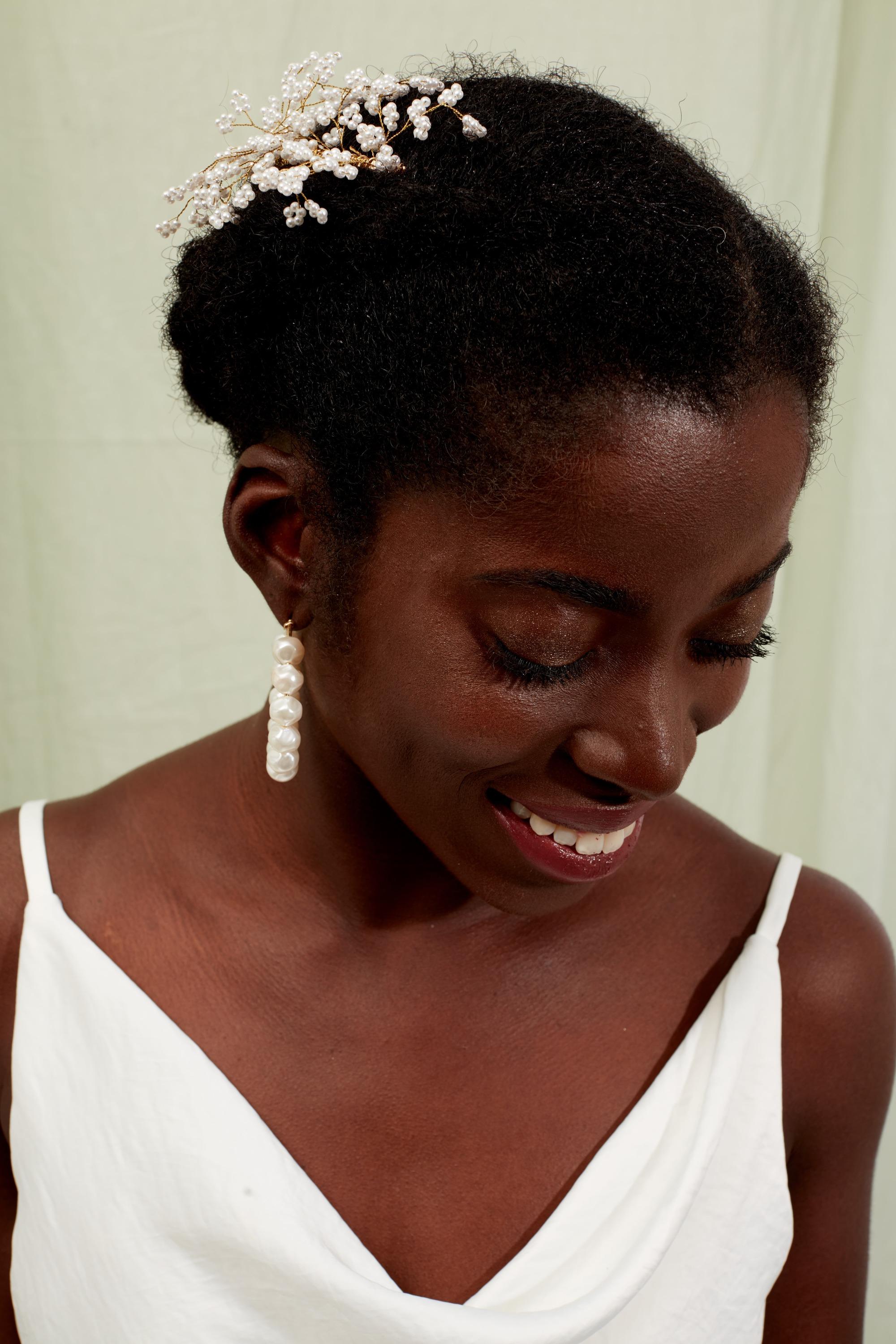 Bridesmaid hairstyles: Woman with natural low braided hairstyle with flowers in it, wearing pearl earrings and white dress in studio