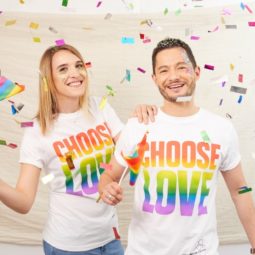 Blonde woman and brown haired man wearing Pride tshirts and waving rainbow flags with confetti