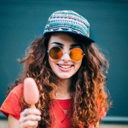 Woman with spiral perm wearing a hat, sunglasses and eating an ice lolly