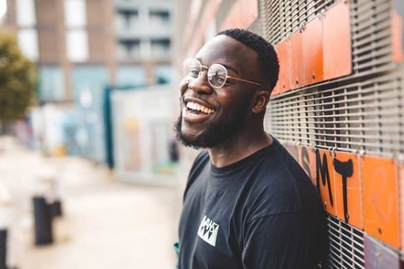Young black man wearing round glasses leaning against a wall