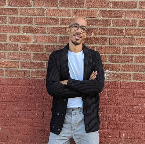 Bald, light skinned black man with glasses leaning against a brick wall
