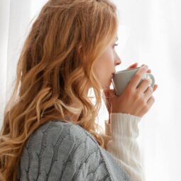 woman sipping from a mug looking through a curtained window
