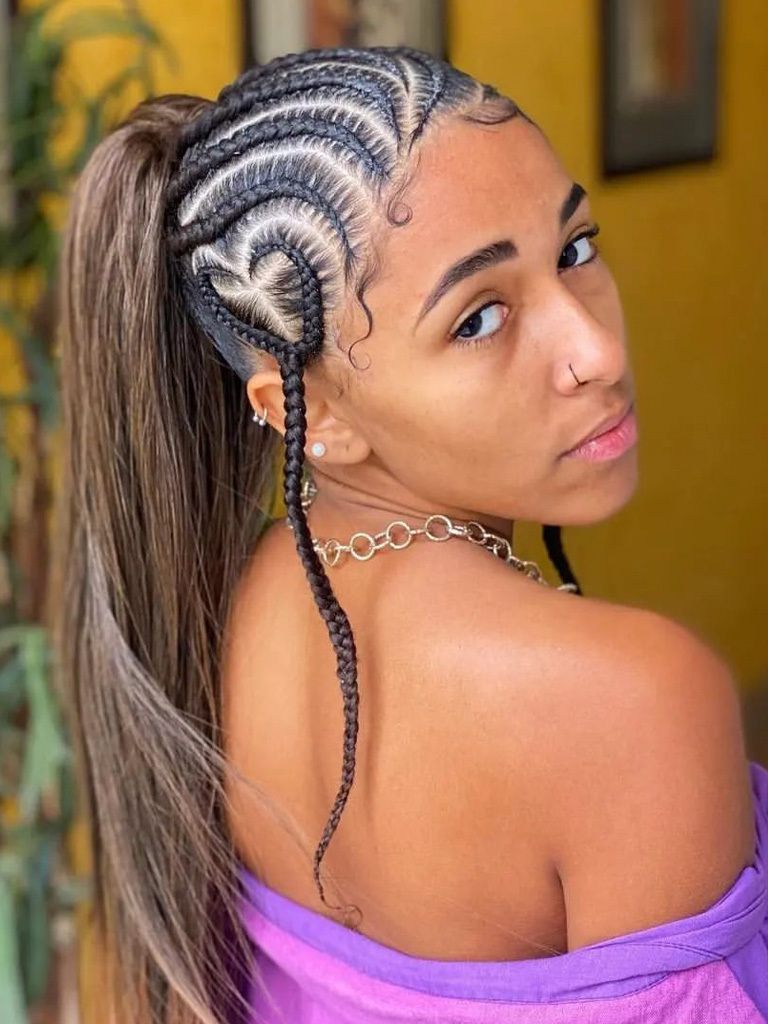 Woman with instricate cornrows in a curving pattern, forming a heart above the ear. Long loose ponytail