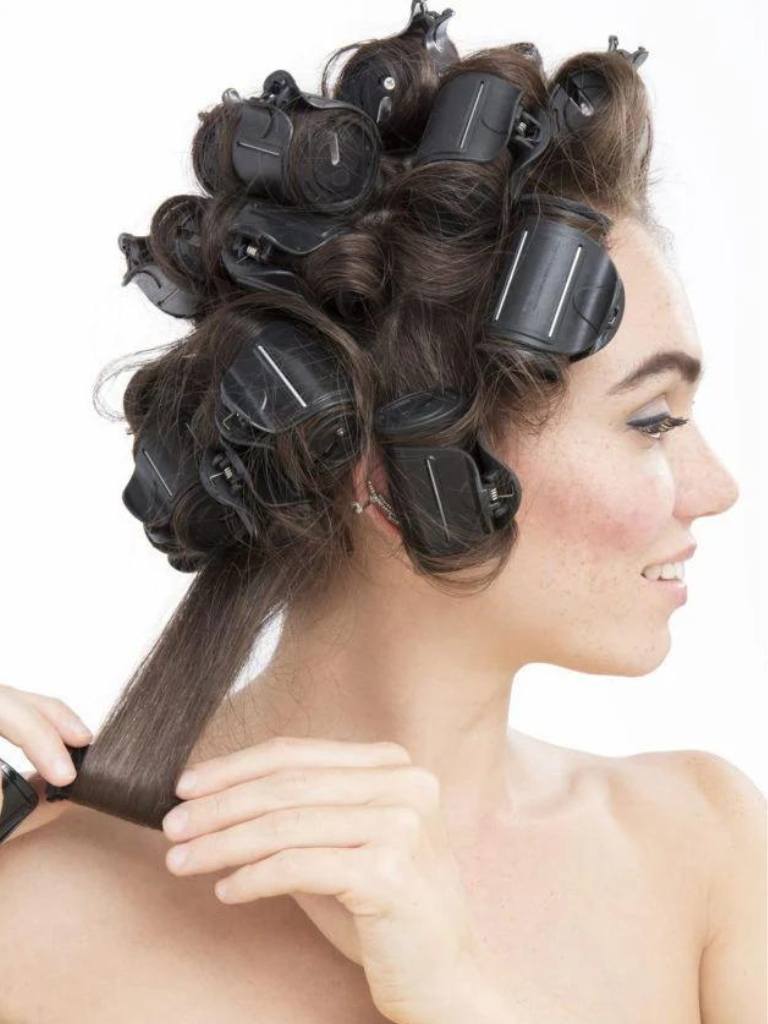 How to use hot rollers: Brunette model with damp long medium brown hair wearing a yellow jumpsuit starting to roll a heated roller