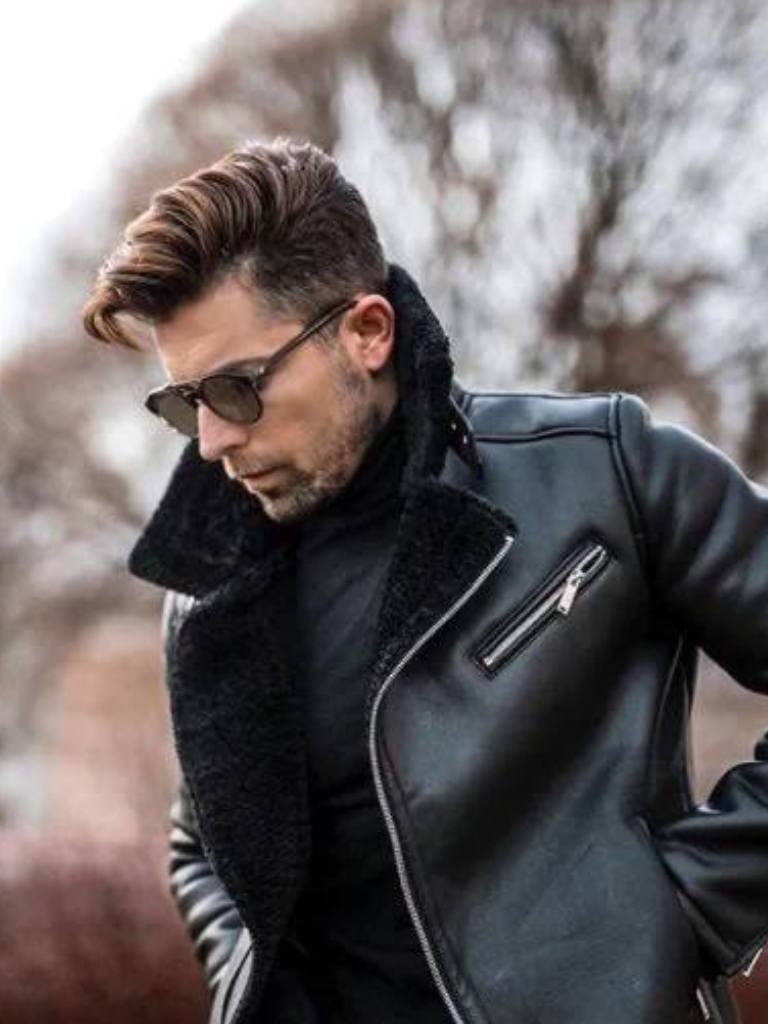 brunette man standing outside in front of a tree wearing a leather jacket and sungalsses with a grown out taper comb over hair cut